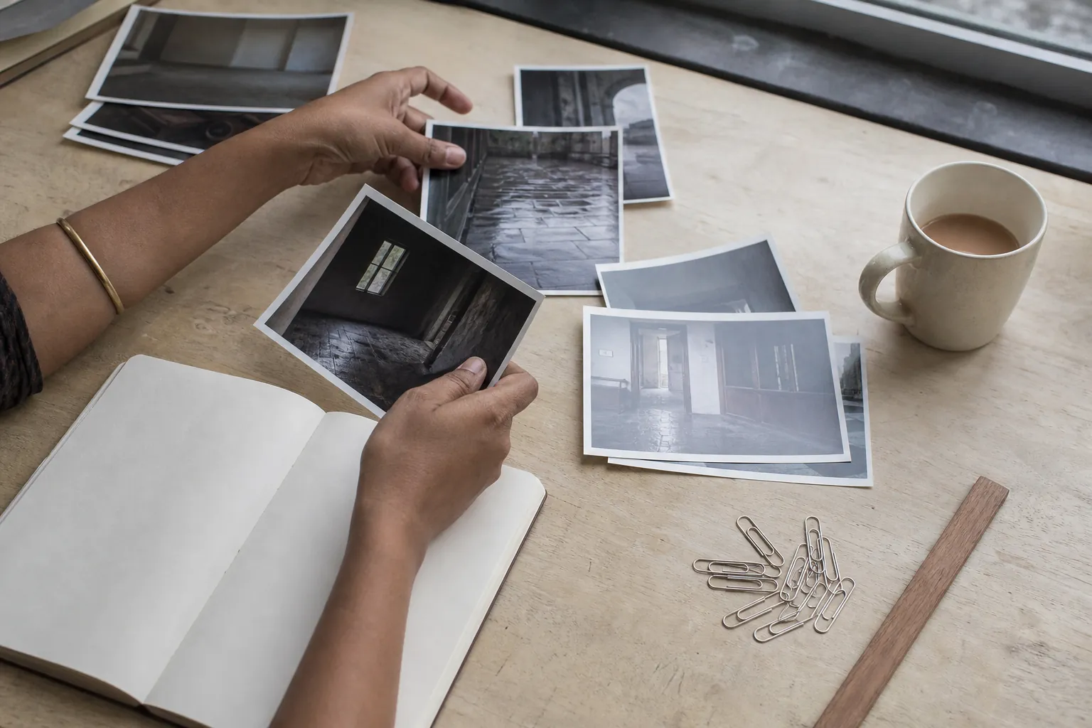 Close view of hands laying photographic prints beside a handwritten notebook, paperclips, a ruler, and a mug of tea on a pale wood desk under a window, 1600x900